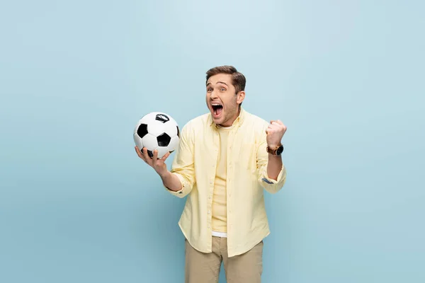 Young amazed man in yellow shirt holding football and cheering on blue — Stock Photo