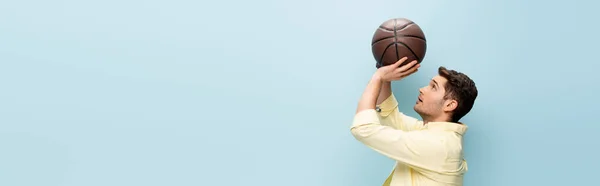 Side view of man in yellow shirt throwing basketball isolated on blue, banner — Stock Photo