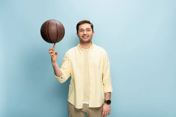 Happy man in yellow shirt spinning basketball on finger on blue — Stock Photo