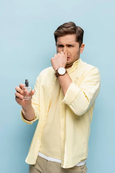 Joven disgustado en camisa amarilla sosteniendo botella con perfume y cubriendo la nariz en azul - foto de stock