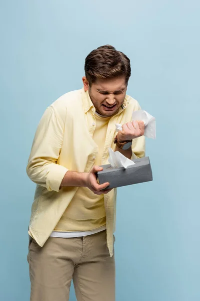Больной человек чихает и держа коробку ткани изолированы на синий — стоковое фото