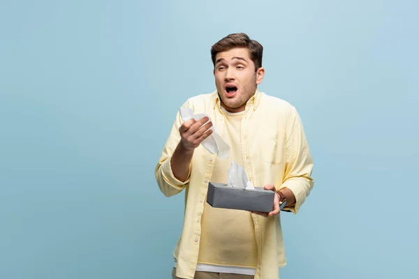 Sick man sneezing and holding tissue box with napkins isolated on blue — Stock Photo