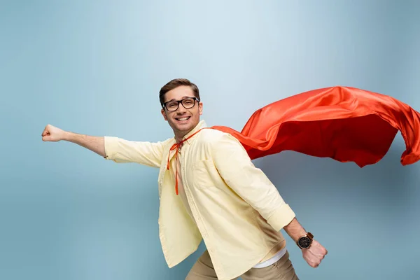 Feliz superhéroe en gafas y capa roja de pie con la mano extendida en azul - foto de stock