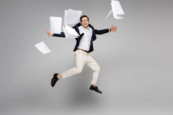 Aufgeregter Geschäftsmann springt in voller Länge in die Nähe von Dokumenten auf grau — Stockfoto