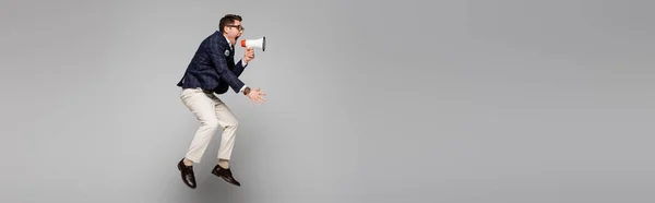 Longitud completa de hombre de negocios saltando y gritando en megáfono en gris, bandera - foto de stock