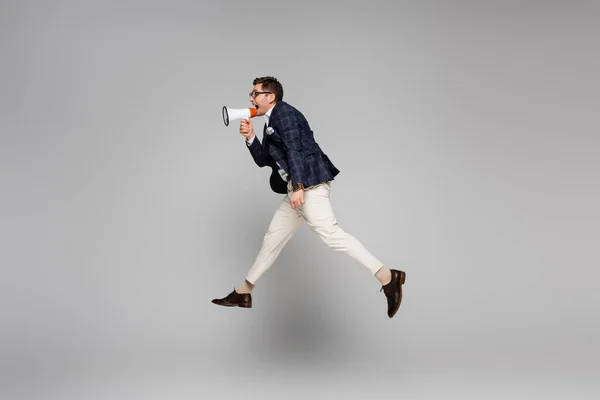 Vista lateral del hombre de negocios saltando y gritando en megáfono sobre gris - foto de stock