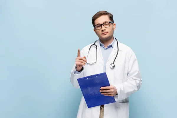 Médico en gafas y abrigo blanco sujetando el portapapeles y mirando a la cámara mientras señala con el dedo en azul - foto de stock
