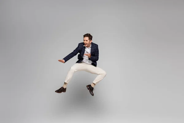 Full length of cheerful young man in blazer levitating on grey — Stock Photo