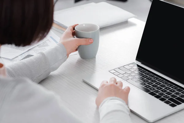 Cropped view of teleworker holding cup near laptop and papers on table — Stock Photo