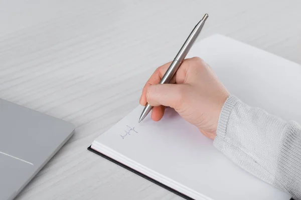 Cropped view of freelancer writing on notebook near laptop on table — Stock Photo