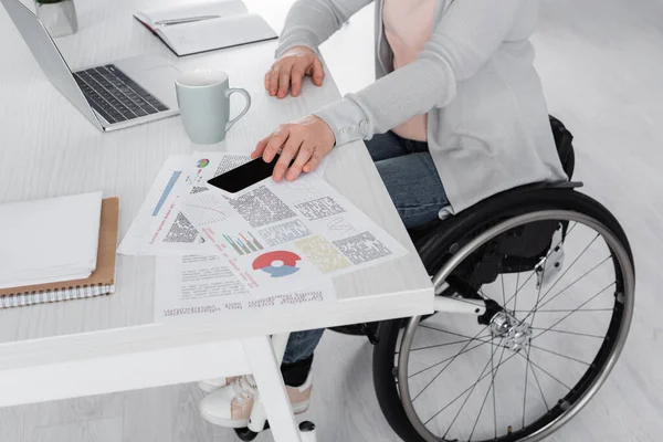 Cropped view of disabled freelancer taking smartphone near laptop and documents on table — Stock Photo