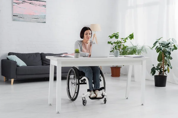 Femme handicapée en fauteuil roulant en utilisant un ordinateur portable près de la tasse et de l'usine — Photo de stock