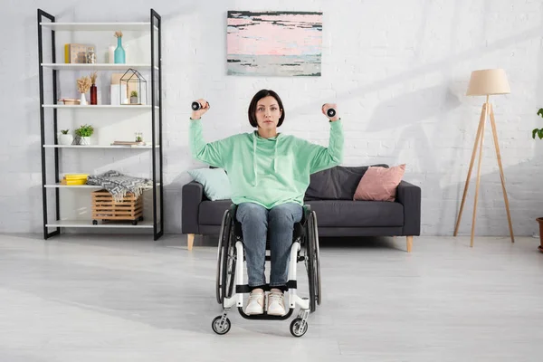Brünette Frau trainiert mit Hanteln im Rollstuhl und schaut zu Hause in die Kamera — Stockfoto