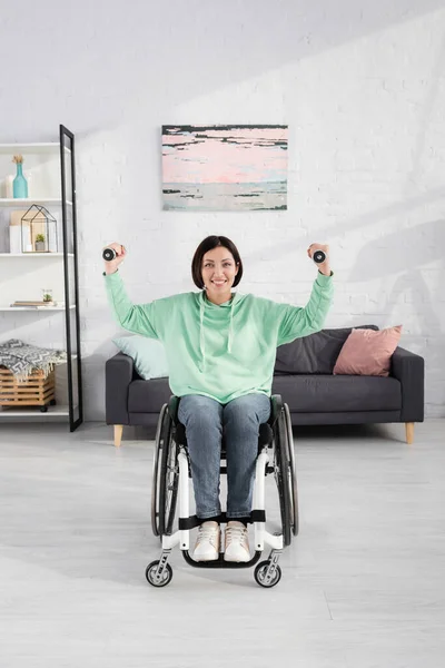 Femme gaie en fauteuil roulant regardant la caméra pendant l'entraînement avec des haltères — Photo de stock