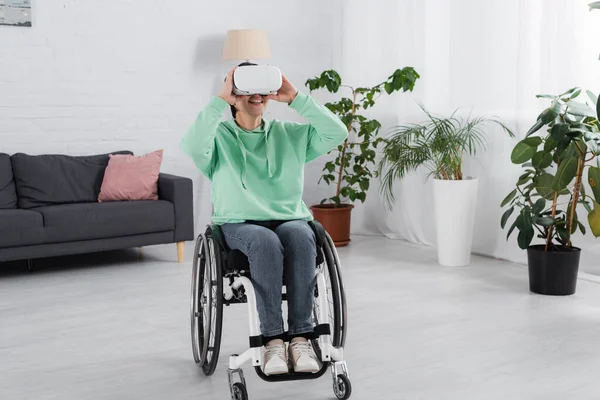 Mujer alegre en silla de ruedas con auriculares vr en la sala de estar - foto de stock