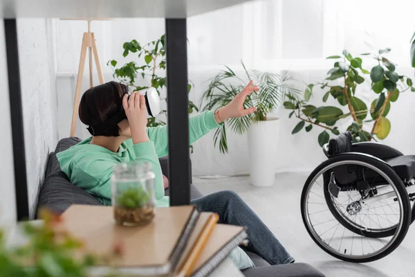 Mujer discapacitada en auriculares vr sentado en el sofá cerca de la silla de ruedas en la sala de estar - foto de stock