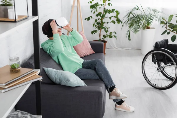 Glücklich unzufriedene Frau in vr Headset sitzt auf Sofa neben Rollstuhl und Schrank auf verschwommenem Vordergrund — Stockfoto