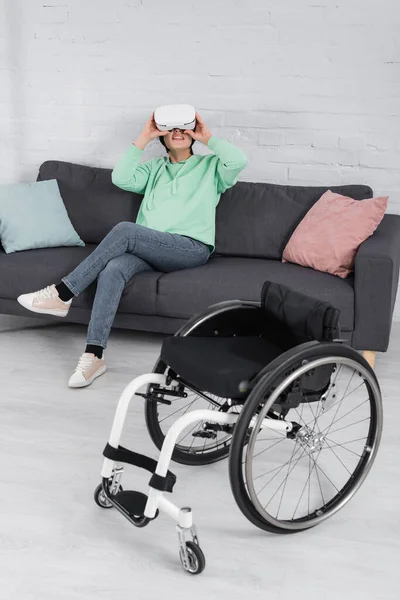 Femme handicapée souriante dans le casque vr assis sur le canapé près du fauteuil roulant — Photo de stock