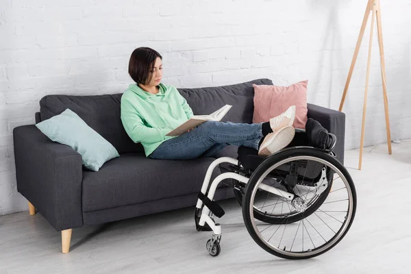 Brünette Frau liest Buch auf Couch neben Rollstuhl — Stockfoto