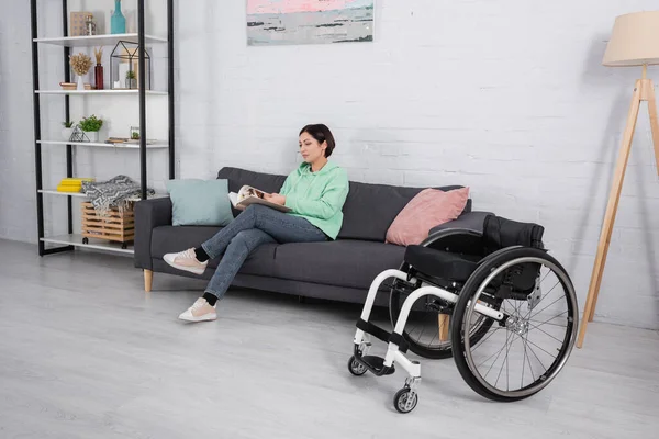 Behinderte Frau liest Buch, während sie auf Couch neben Rollstuhl sitzt — Stockfoto