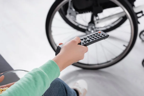 Vue recadrée de la femme tenant télécommande et pop-corn près de fauteuil roulant sur fond flou — Photo de stock