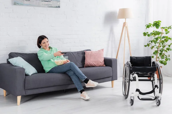Femme gaie avec télécommande et pop-corn regarder la télévision près de fauteuil roulant — Photo de stock