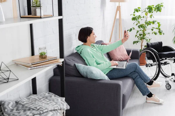 Lächelnde Frau mit Smartphone und Laptop in der Nähe von Rollstuhl — Stockfoto