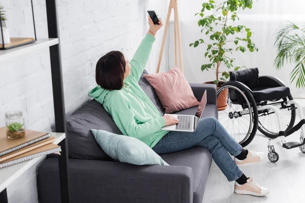 Behinderte Frau mit Laptop macht Selfie auf Smartphone neben Rollstuhl zu Hause — Stockfoto