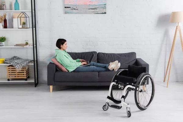 Vue latérale d'une femme mécontente utilisant un ordinateur portable sur un canapé près d'un fauteuil roulant — Photo de stock