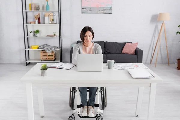 Handicapped freelancer utilizzando laptop vicino a documenti con grafici e tazza sul tavolo — Foto stock
