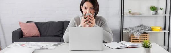 Mujer morena bebiendo café y mirando a la cámara cerca de dispositivos y papeles con gráficos en la mesa, pancarta - foto de stock