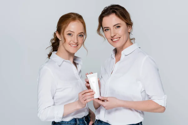 Mulheres sorridentes em camisas brancas segurando tubo com creme isolado em cinza — Fotografia de Stock