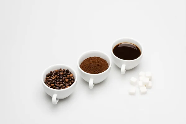 Xícaras com café preparado e moído perto de grãos e cubos de açúcar em branco — Fotografia de Stock