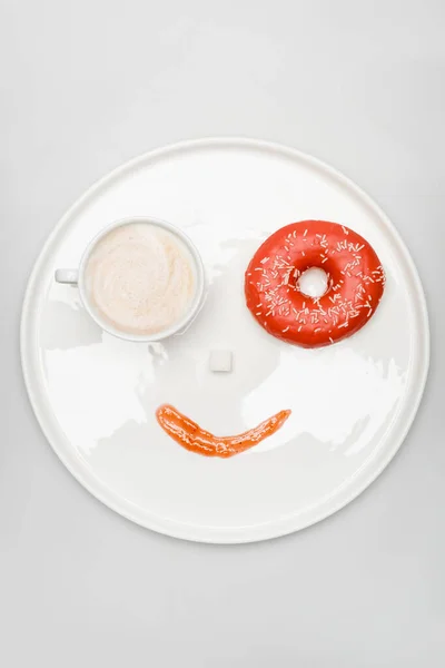 Vista superior de la cara sonriente hecha de latte en taza, donut, cubo de azúcar y mermelada en bandeja redonda en blanco — Stock Photo