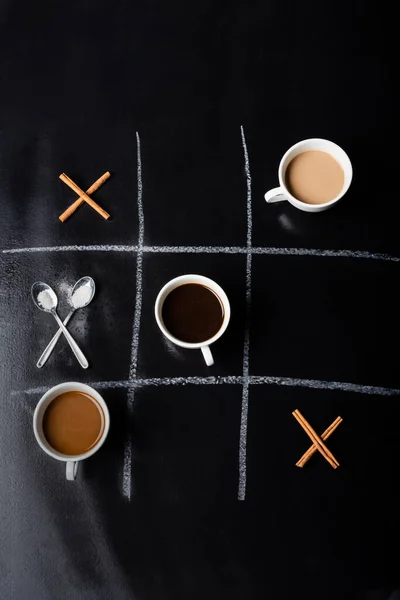 Top view of tic tac toe game with spoons, cinnamon sticks and cups of coffee on black — Stock Photo