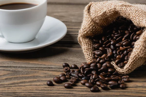 Sacchetto di sacco con chicchi di caffè torrefatto vicino tazza su superficie di legno — Foto stock