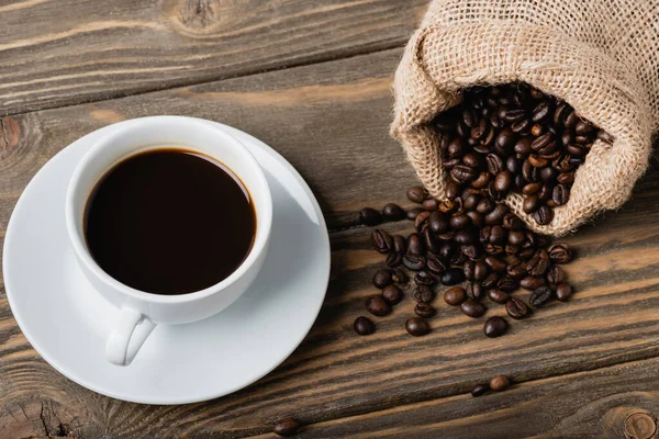 Vista superior del saco con granos de café tostados cerca de la taza en la superficie de madera — Stock Photo