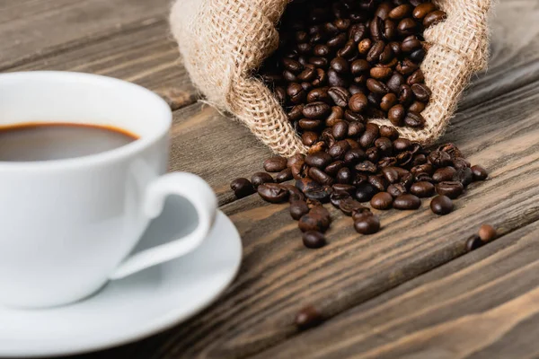 Мішок з смаженими кавовими зернами поблизу розмитої чашки на дерев'яній поверхні — стокове фото