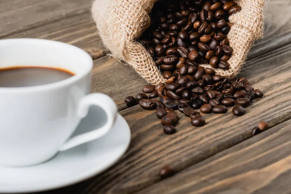 Saco saco com grãos de café torrados perto copo borrado na superfície de madeira — Fotografia de Stock