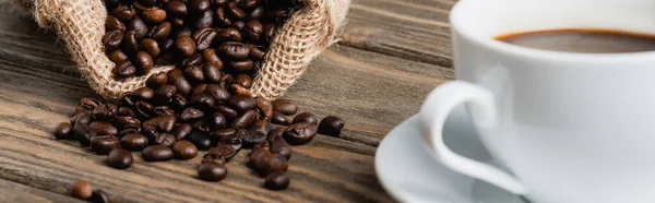 Sac à dos avec grains de café torréfiés près de tasse floue sur la surface en bois, bannière — Photo de stock