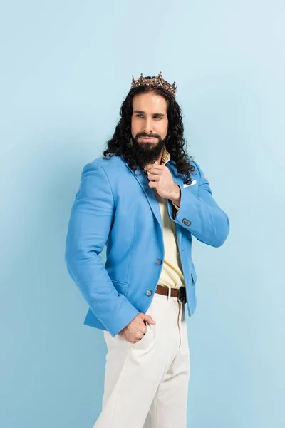 Homme hispanique barbu en couronne et veste posant avec la main dans la poche isolé sur bleu — Photo de stock