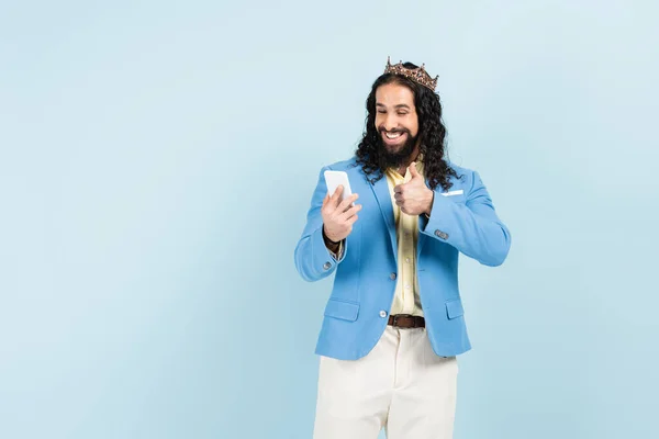 Hombre hispano barbudo con chaqueta y corona mostrando el pulgar hacia arriba durante una videollamada aislada en azul — Stock Photo