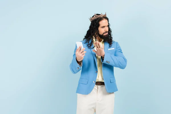 Beleidigter hispanischer Mann in Jacke und Krone zeigt Stopp-Geste, während er Smartphone isoliert auf blau hält — Stockfoto