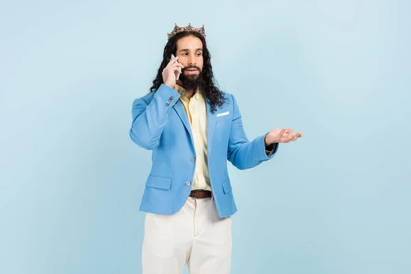 Homme hispanique barbu en veste et couronne parler sur smartphone isolé sur bleu — Photo de stock