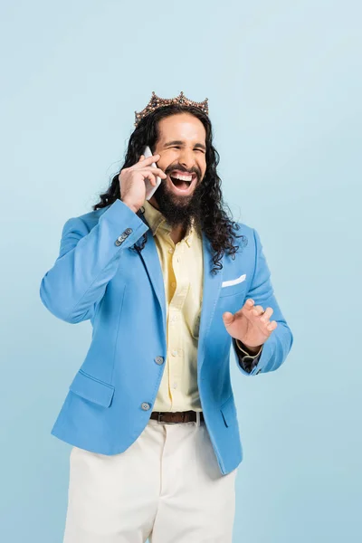 Heureux hispanique homme en veste et couronne parler sur smartphone et rire isolé sur bleu — Photo de stock