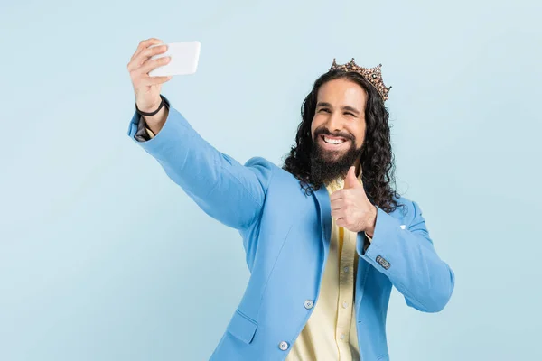 Homem hispânico feliz em casaco e coroa mostrando polegar para cima e tomando selfie isolado em azul — Fotografia de Stock