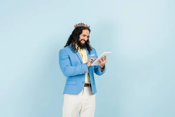 Hombre hispano feliz con chaqueta y corona sosteniendo tableta digital aislada en azul — Stock Photo