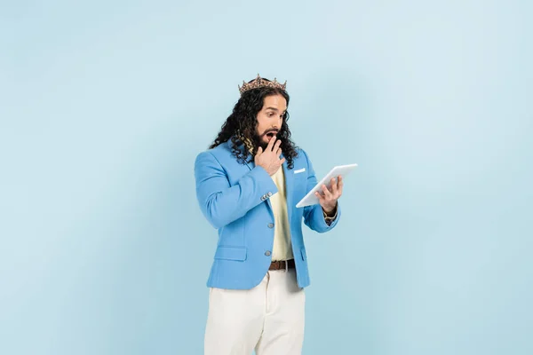 Sorprendido hombre hispano con chaqueta y corona sosteniendo tableta digital aislado en azul — Stock Photo