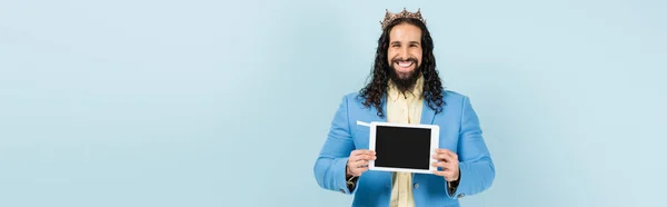 Homem hispânico feliz em casaco e coroa segurando tablet digital com tela em branco isolado em azul, banner — Fotografia de Stock