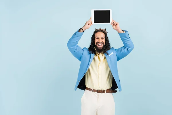 Glücklicher hispanischer Mann in Jacke und Krone mit digitalem Tablet und leerem Bildschirm über dem Kopf isoliert auf blau — Stockfoto